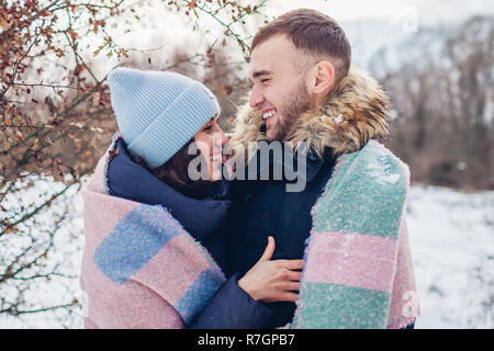 Beautiful loving couple hugging and laughing in winter forest. Happy people warming covered with blanket Stock Photo