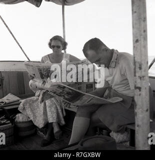 1950s, a man and wpman sitting on a boat on Lake Victoria, Uganda, reading a newspaper, The Daily Mail. The British daily newspaper in this era was a broadsheet and would be sent to British colonies and protectorates overseas, such as Uganda was, before the country became independent in 1962. Stock Photo