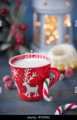 Closeup on red cup of milk with Christmas deer design, bundt cake, Xmas lights in lantern and berry decorations Stock Photo