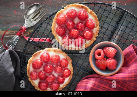 Red raspberry shortbread tarts with vanille custard and glazed fresh raspberries on cooling rack on dark brown wood, top view Stock Photo