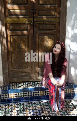 A long haired brunette hippie girl wearing pink, red and flowers sits in a tiled doorway in Morocco Stock Photo