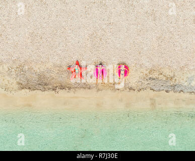 Aerial  view of three women lying on big inflatable star, donut, flamingo shaped mattresses on beach. Stock Photo