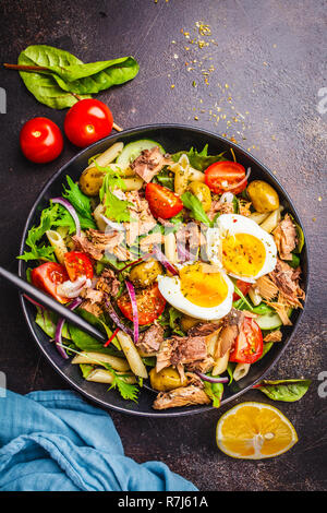 Tuna salad with pasta, olives, vegetables and poached egg in a black plate on a dark background, top view. Stock Photo