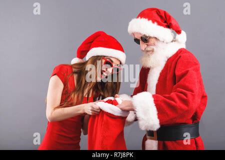 Santa Claus with white beard wearing sungasses and young mrs. Claus wearing Santa hat, red dress and sunglasses standing on the gray background, mrs. Santa searching some presents in the Santa Claus bag, New Year, Christmas, holidays, souvenirs, gifts, shopping, discounts, shops, Snow Maiden Santa Claus,make-up, hairstyle, carnival. Stock Photo