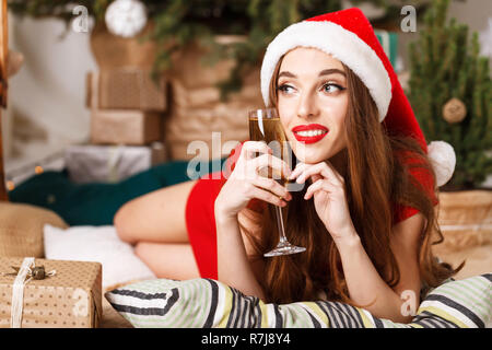 Hilarious christmas woman wearing Santa hat and red dress laying on the floor near the new year spruce with glass of champagne, New Year, Christmas, holidays, souvenirs, gifts, shopping, discounts, shops, Snow Maiden Santa Claus,make-up, hairstyle, carnival. Stock Photo