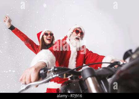 Santa Claus with white beard wearing sungasses and young mrs. Claus wearing Santa hat, red sweater and sunglasses rejoicing in the snow while riding a motorcycle when snowing, New Year, Christmas, holidays, souvenirs, gifts, shopping, discounts, shops, Snow Maiden Santa Claus,make-up, hairstyle, carnival. Stock Photo