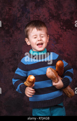 Cute kid holding French bread against dark red background. Little boy eating the french baguette Stock Photo