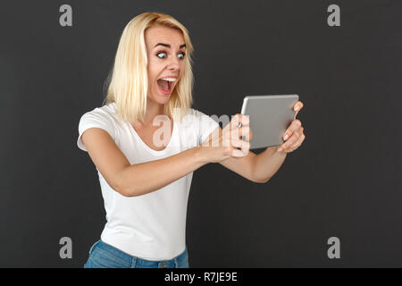 Freestyle. Woman standing isolated on black wall watching video on digital tablet looking at screen surprised Stock Photo