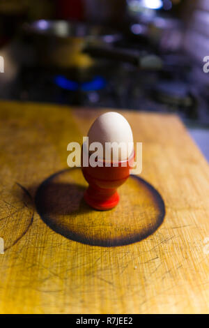 boiled egg in red ceramic egg cup sitting within burnt circle on bamboo chopping board with gas hob and pot in background in domestic kitchen Stock Photo
