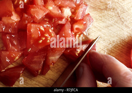 Overhead shot of hand holding knife to chop and slice juicy, red raw salad tomatoes on wooden chopping board. Stock Photo