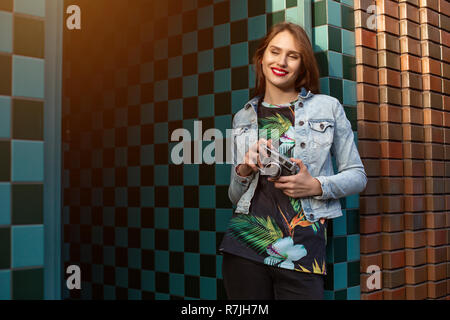 Cool funny girl model with retro film camera wearing a denim jacket, dark hair outdoors over city wall in a cage background. Sun flare Stock Photo