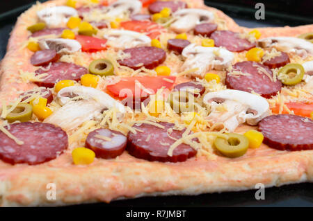 Delicious pizza in a heart shape, ready for baking. Pizza with mushrooms, salami, pepperoni, olives, corn on a baking tray. Stock Photo