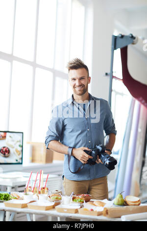 Waist up portrait of smiling photographer looking at camera while taking images of party table in studio Stock Photo