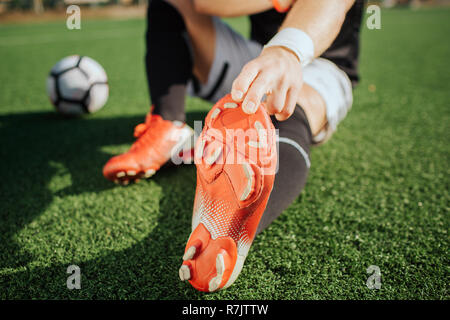Cut view of player sitting on lawn outside and stretching. He reaches to feet. Ball lying besides him. It is sunny weather. Stock Photo
