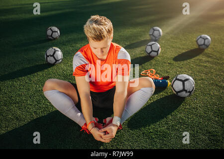 Young football player sitting on lawn and stretching. He keeps feet together. Sun shining outside. Balls lying behind him. Stock Photo