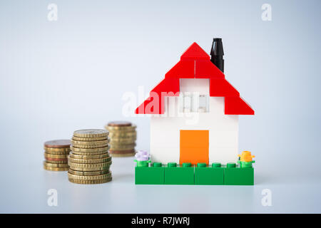 Stacks of coins and simple house made of toy building bricks, on gray background. Concept photo of mortgage, real estate business or house ownership. Stock Photo