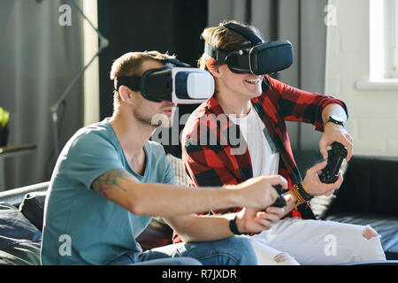 Two young men sitting on the couch and playing game with virtual reality headset and joypad Stock Photo