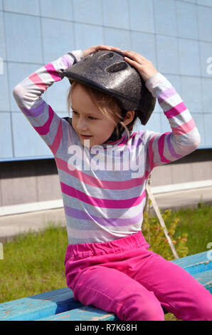 Blonde girl sits in pink jeans on a bench on a bright summer day in a roller helmet in a striped puller with arms raised. Photo Stock Photo