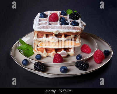 A stack of four home made Belgian waffles dusted with sugar, with fresh berries and mint leaves, on a beige platter, set on dark brown background Stock Photo