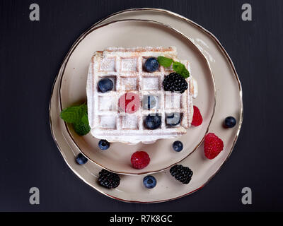 A stack of four home made Belgian waffles dusted with sugar, with fresh berries and mint leaves, on a beige platter, set on dark brown background, fla Stock Photo