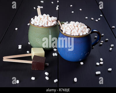 Two cups with hot chocolate and mini marshmallows, one cup being blue and the other one green, with white and dark chocolate cubes, on dark brown back Stock Photo