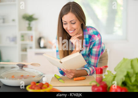 Beautiful young woman cooking healthy meal in the domestic kitchen. She is reading recipe in the cookbook. Stock Photo