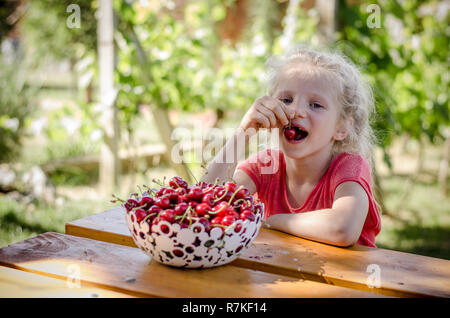 adorable blond girl eating cherry fruit in the garden Stock Photo