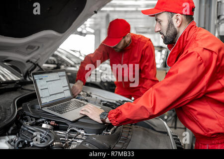Two auto mechanics in red uniform doing engine diagnostics with computer in the car service Stock Photo