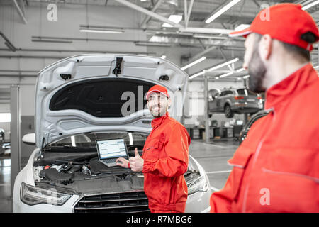 Two auto mechanics in red uniform doing engine diagnostics with computer in the car service Stock Photo