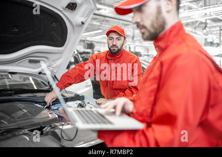 Two auto mechanics in red uniform doing engine diagnostics with computer in the car service Stock Photo