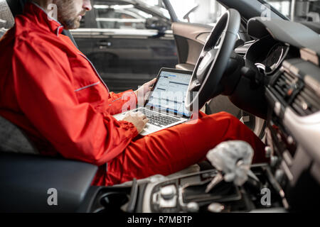 Auto mechanic in red uniform diagnosing car with computer sitting on the driver seat at the car service Stock Photo