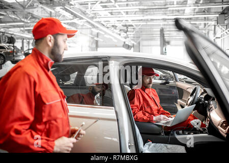 Two male auto mechanics in red uniform diagnosing car with computer at the car service Stock Photo