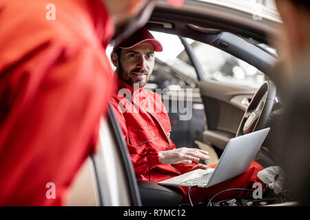 Auto mechanic in red uniform diagnosing car with computer sitting on the driver seat at the car service Stock Photo