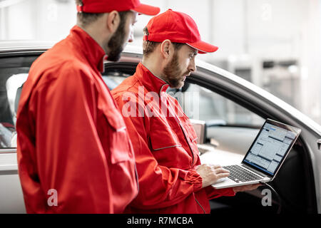 Two male auto mechanics in red uniform diagnosing car with computer standing near the luxury car at the workshop Stock Photo