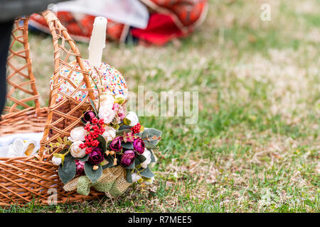 Eggs and Kulich Russian Orthodox Easter blessing wicker straw basket closeup, candle on grass ground outside at church, flowers Stock Photo