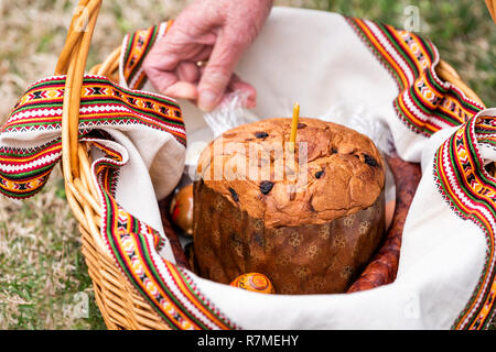 Eggs and raisin Kulich Russian Orthodox Easter blessing wicker straw basket, candle on grass ground outside at church, hands Stock Photo
