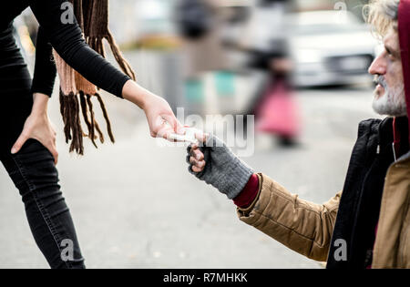 A midsection of woman giving money to homeless beggar man sitting in city. Stock Photo