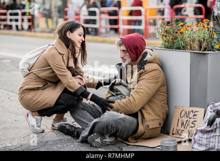 Young woman giving money to homeless beggar man sitting in city. Stock Photo