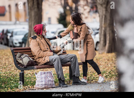 Young woman giving food to homeless beggar man sitting on a bench in city. Stock Photo