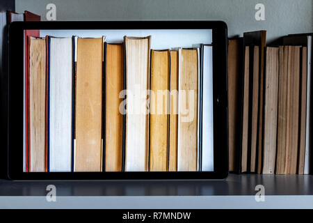 Tablet and books on a bookshelf. The bright screen shows the books behind. Dark background. Stock Photo