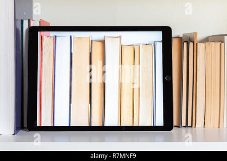 Tablet and books on a bookshelf. The screen shows the books behind. Light background. Stock Photo