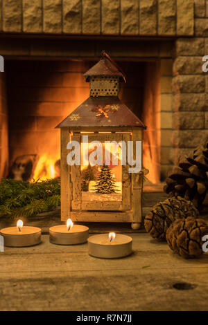 Merry Christmas before cozy fireplace with christmas lantern ,candles and cones , on old brown table, vertical orientation. Stock Photo