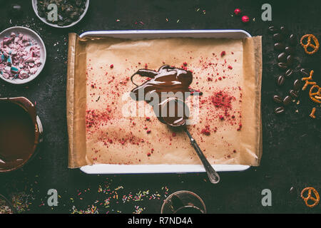 Spoon with melted chocolate on tray with red dried fruits powder topping on dark kitchen table background with various flavoring ingredients for choco Stock Photo