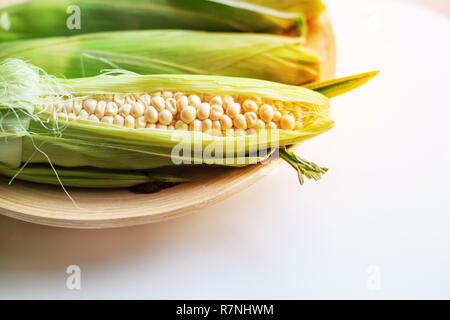 fresh ears of white milky sweet corns with green leaves on wooden tray with white table background. Organic vegetable, vegan farm food, food nutrition Stock Photo