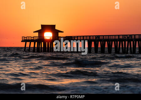 Naples, Florida vibrant orange sunset, in gulf of Mexico with sun peeking in behind Pier wooden jetty, with horizon and dark silhouette ocean waves Stock Photo