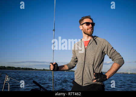 Confident and happy sailor stand on yacht. He looks away. Guy holds the rope. He is ready for a trip. Sun goes down. Stock Photo