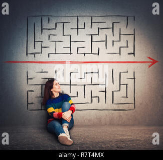 Casual young woman sitting on the floor finding a solution to solve problem and escape from labyrinth. Breaking the rules, as a red arrow pierce the m Stock Photo