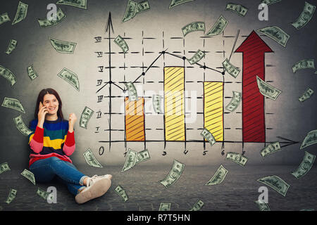 Pretty woman sitting on the floor talking on phone holding fist up as win symbol, positive emotion, good news listening. Imagining a rising graph of v Stock Photo