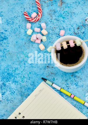 Cup with coffee and marshmallows, blank open notebook with pen on abstract blue background. Top view. Flat mockup. space for text. Stock Photo