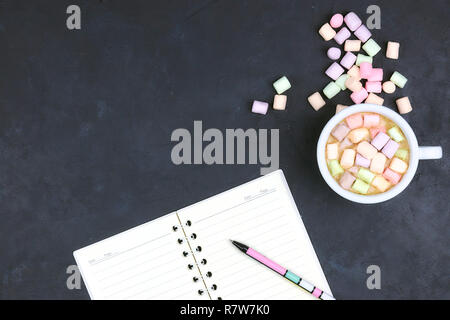 Cup with coffee and marshmallows, blank open notebook with pen on black concrete background. View from above. Flat layout. space for text. hot drink.
